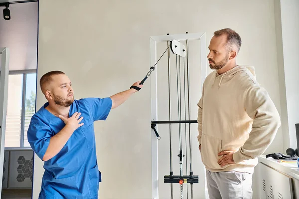 Joven fisioterapeuta en uniforme azul instruyendo al hombre cerca de la máquina de ejercicio en el centro kinesio - foto de stock