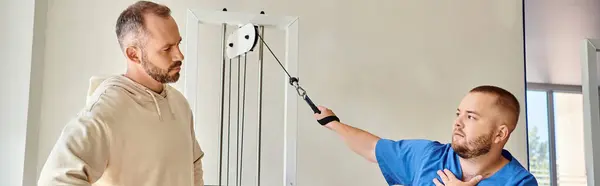 Joven médico en uniforme azul instruyendo al hombre cerca de la máquina de entrenamiento en el centro de kinesio, pancarta - foto de stock