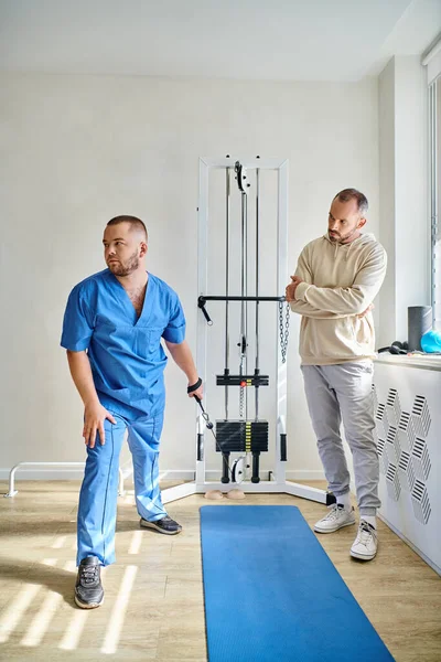 Recovery specialist in blue uniform instructing man near training machine in kinesio center — Stock Photo