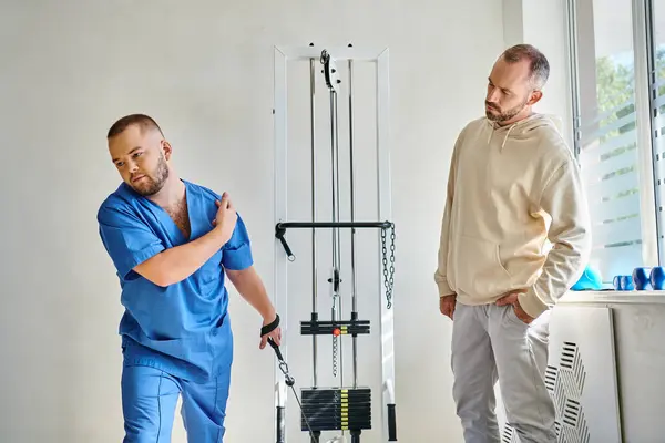 Physiothérapeute en uniforme bleu instruisant l'homme près de la machine d'entraînement dans le centre de réadaptation — Photo de stock