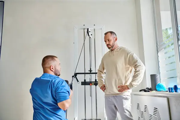 Rehabilitólogo experimentado que trabaja en la máquina del ejercicio, instrucción del hombre en el centro del kinesio - foto de stock