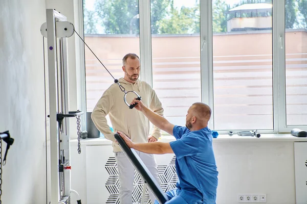 Entrenamiento profesional del rehabilitólogo en la máquina del ejercicio, instrucción del hombre en el centro del kinesio - foto de stock