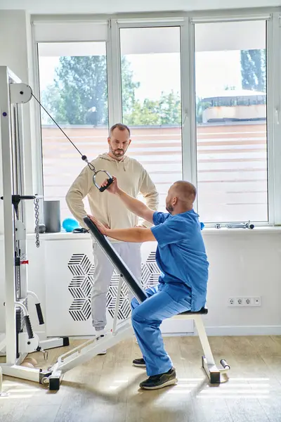 Médico en el entrenamiento uniforme azul en la máquina de ejercicio, instrucción del hombre en el centro moderno de kinesio - foto de stock
