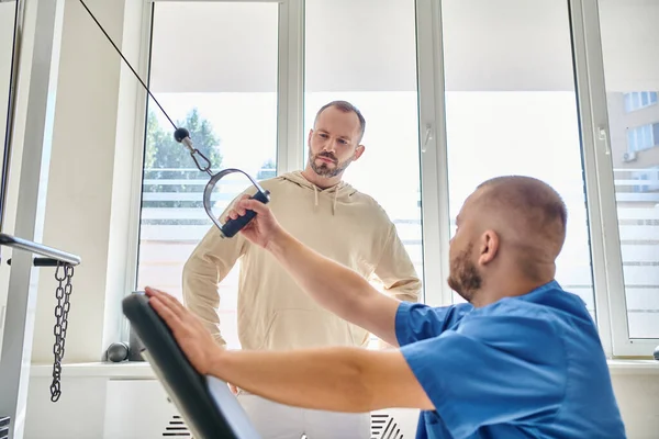 Homem olhando para o treinamento fisioterapeuta profissional na máquina de exercício no centro de cinesiologia — Fotografia de Stock