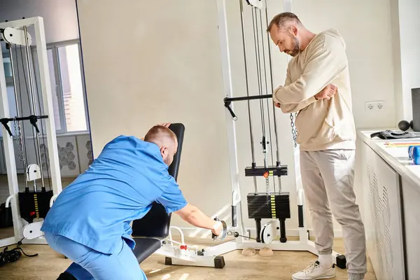 Paciente masculino que mira el entrenamiento del rehabilitólogo joven en la máquina del ejercicio en el centro de kinesiología - foto de stock