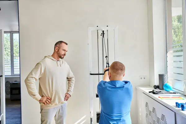 Man looking at recovery expert in blue uniform training on exercise machine in kinesiology center — Stock Photo