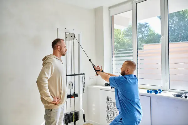 Young rehabilitologist showing male patient how to work out on exercise machine in kinesio center — Stock Photo