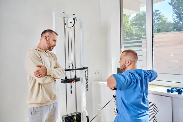 Hombre con los brazos cruzados mirando el entrenamiento del rehabillitologist en la máquina del ejercicio en el centro de recuperación - foto de stock