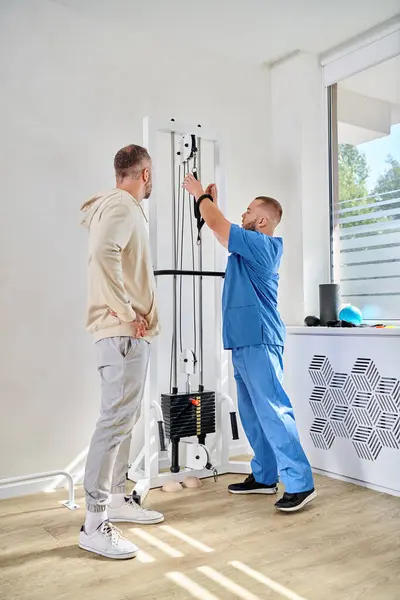 Молодий лікар у синій формі, що показує тренувальне обладнання для пацієнта чоловічої статі в центрі відновлення кінезіо — стокове фото