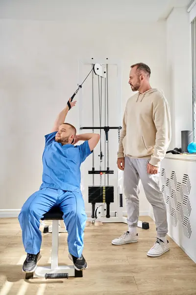 Entrenamiento de fisioterapeuta joven en la máquina de ejercicio durante la consulta del hombre en el centro de kinesio - foto de stock
