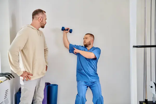 Man looking at young rehabilitologist showing how to train arm with dumbbell in kinesiology center — Stock Photo
