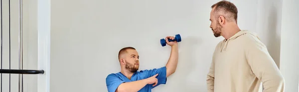 Young rehabilitologist showing how to train arm with dumbbell to man in kinesio center, banner — Stock Photo