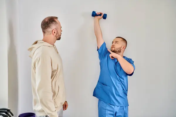 Кваліфікований реабілітолог, який показує вправи з гантелі до свого пацієнта в центрі відновлення кінезіо — стокове фото