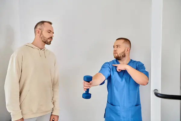 Young doctor in blue uniform showing how to train with dumbbell to man in recovery kinesio center — Stock Photo