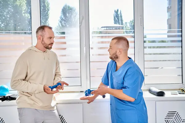 Young rehabilitologist and bearded man holding dumbbells and talking in recovery kinesio center — Stock Photo