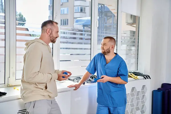 Jeune spécialiste en uniforme bleu parlant à son patient masculin dans le centre de kinesio, réadaptation — Photo de stock