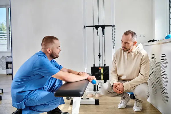 Riabilitologo e paziente di sesso maschile discutere piano di trattamento vicino macchina esercizio nel centro di Kinesio — Foto stock