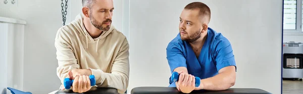 Physician in blue uniform and his male patient exercising with dumbbells in kinesio center, banner — Stock Photo