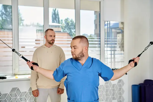 Skilled doctor in blue uniform working out on exercise machine instructing man in kinesio center — Stock Photo
