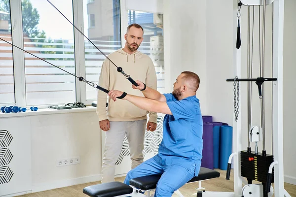 Rehabilitologist in blue uniform showing exercise on training machine to man in kinesio center — Stock Photo