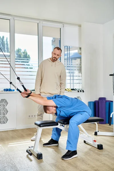 Médico profesional que muestra el ejercicio en la máquina de entrenamiento al hombre en el gimnasio del centro kinesio - foto de stock