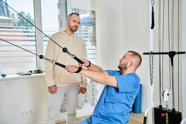 Joven médico experimentado que muestra el ejercicio en la máquina de entrenamiento al hombre en el gimnasio del centro kinesio - foto de stock