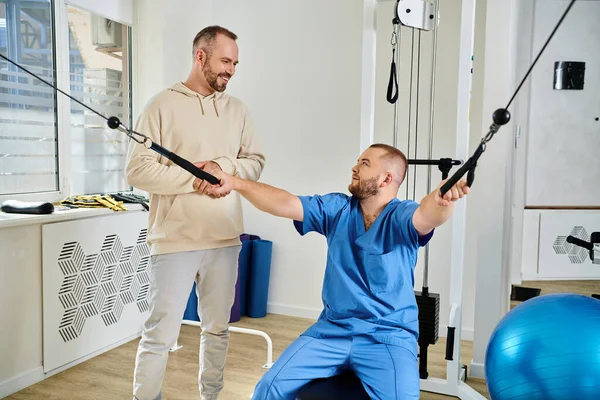 Médico de uniforme azul mostrando exercício na máquina de treinamento para homem sorridente no centro de cinesiologia — Fotografia de Stock
