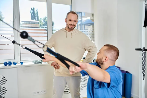 Junge Physiotherapeutin zeigt fröhlichem Mann im Kinesiologie-Zentrum Übung an Trainingsgerät — Stockfoto