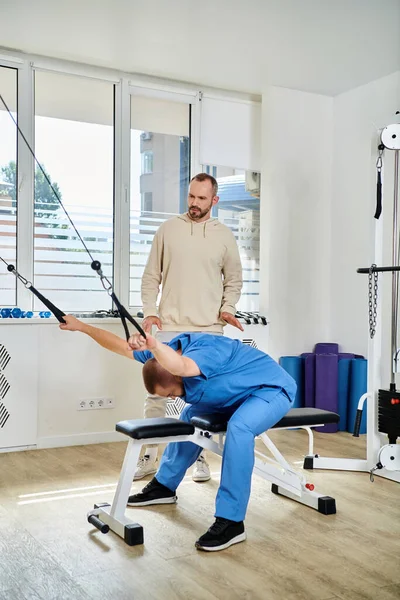 Experienced rehabilitologist showing exercise on training machine to man in kinesiology center — Stock Photo