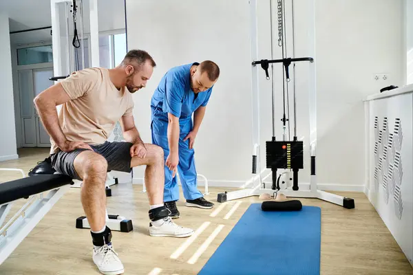 Young doctor pointing at leg belts of man sitting near exercise machine in gym of kinesio center — Stock Photo