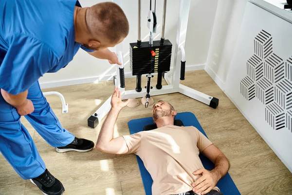Vista ad alto angolo dell'istruttore che assiste il suo paziente sdraiato sul fitness man vicino all'attrezzo da ginnastica — Foto stock