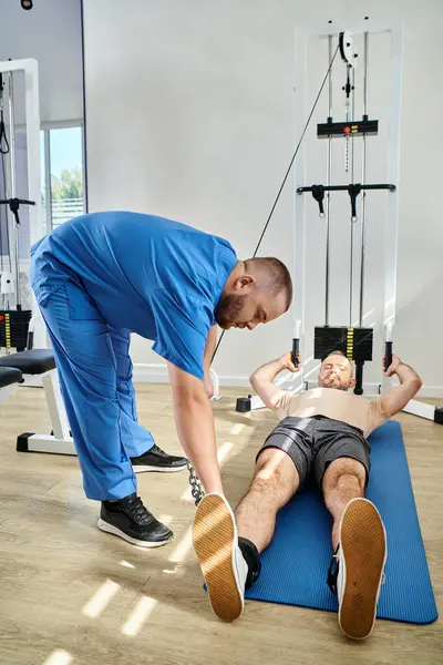 Medico in uniforme blu che assiste l'uomo in abbigliamento sportivo sdraiato sul tappeto fitness vicino alla macchina da ginnastica — Foto stock