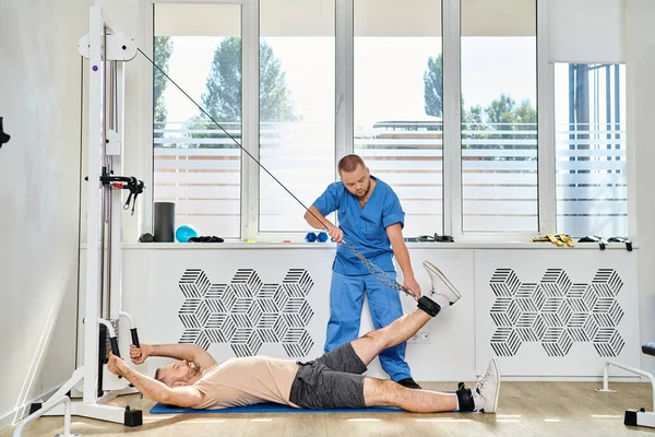 Habilidoso rehabilitólogo que ayuda al hombre a ejercitarse en la máquina de ejercicio en el gimnasio del centro kinesio - foto de stock