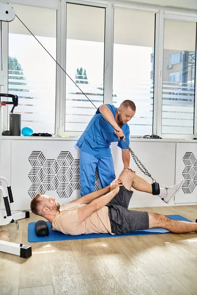 Joven especialista en recuperación que ayuda al hombre a ejercitarse en la máquina de ejercicio en el gimnasio del centro kinesio - foto de stock