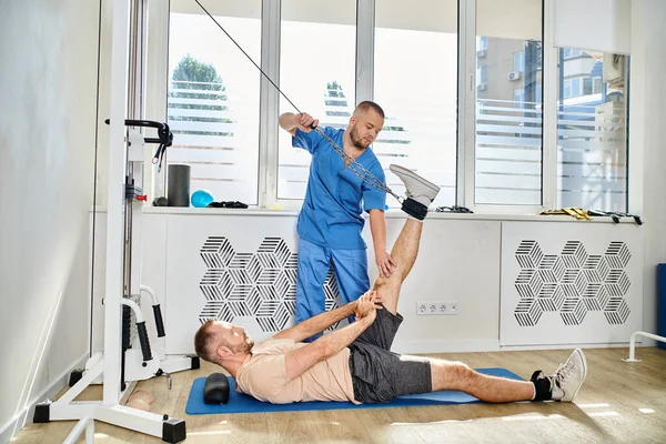 Erfahrener Recovery-Experte unterstützt Mann beim Training an Trainingsgerät im Fitness-Studio des Kinesio-Zentrums — Stockfoto