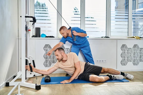 Junger Recovery-Experte unterstützt Mann beim Training an Trainingsgerät im Fitnessstudio des Kinesio-Zentrums — Stockfoto