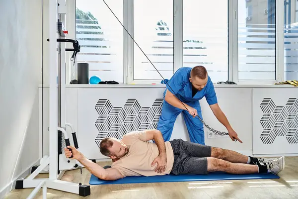 Junge Ärztin in blauer Uniform unterstützt Mann beim Training an Trainingsgerät in Turnhalle des Kinesio-Zentrums — Stockfoto