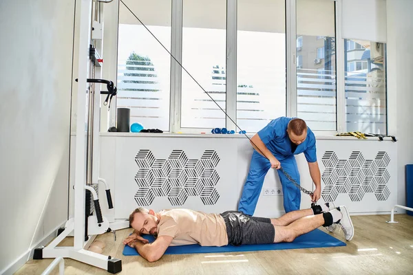 Médico en uniforme azul ayudando al hombre a hacer ejercicio en la máquina de ejercicio en el gimnasio del centro kinesio - foto de stock