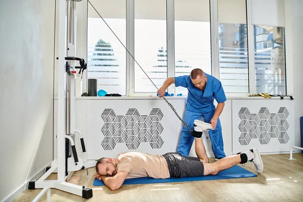 Physician in blue uniform assisting man working out on exercise machine in modern kinesio center — Stock Photo