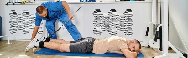 Молодой реабилитолог помогает человеку тренироваться на тренажере в современном кинезио-центре, баннер — стоковое фото