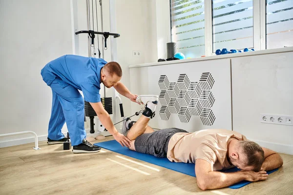 Медичний фахівець в блакитній формі готує чоловіка для відновлення тренувань в тренажерному залі центру кінезіо — стокове фото