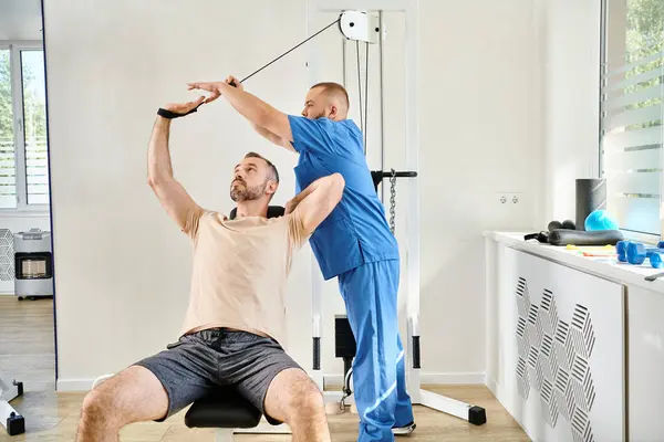 Junger Ausbilder unterstützt Mann beim Erholungstraining an Trainingsgerät im Kinesio-Zentrum — Stockfoto