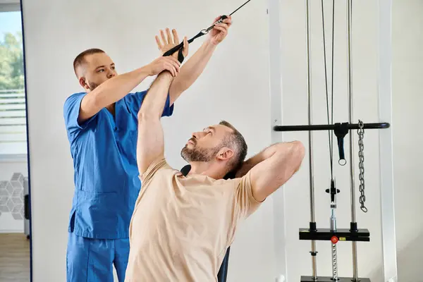 Médico experto que ayuda al hombre durante el entrenamiento de recuperación en la máquina de ejercicio en el centro de kinesio - foto de stock