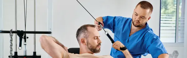 Assistente medico che aiuta l'uomo con l'addestramento di recupero sulla macchina di esercitazione nel centro di kinesio, bandiera — Foto stock