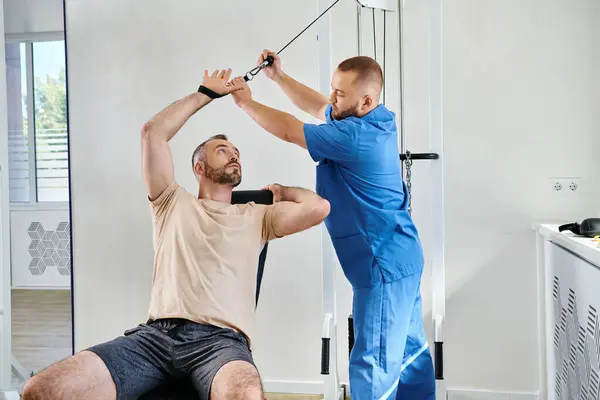 Médico en uniforme azul ayudando al hombre con el entrenamiento de recuperación en la máquina de ejercicio en el centro kinesio - foto de stock