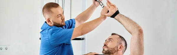 Rehabilitólogo ayudando al hombre con el entrenamiento de recuperación en la máquina de ejercicio en el centro de kinesio, pancarta - foto de stock