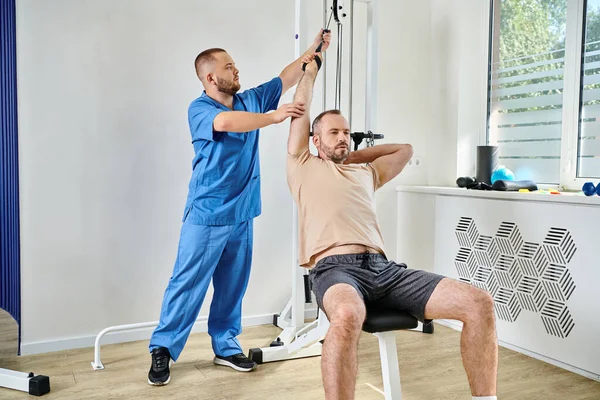 Assistente professionale che sostiene l'uomo che lavora su macchina di esercizio in palestra di centro di kinesio — Foto stock