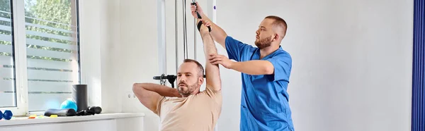 Doctor in blue uniform assisting man working out on exercise machine in kinesio center, banner — Stock Photo