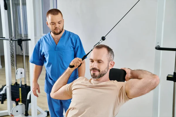 Young instructor assisting smiling man training on exercise machine in gym of kinesio center — Stock Photo