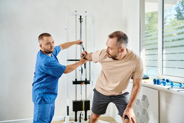 Man in sportswear working out on exercise machine with help of recovery specialist in kinesio center — Stock Photo